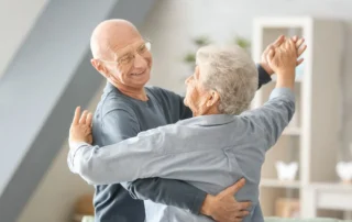 Two elderly people dancing together in a cozy living room, smiling as they hold each other.
